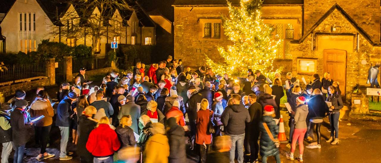 A large group of people sing carols around a lit Christmas tree outside at night.