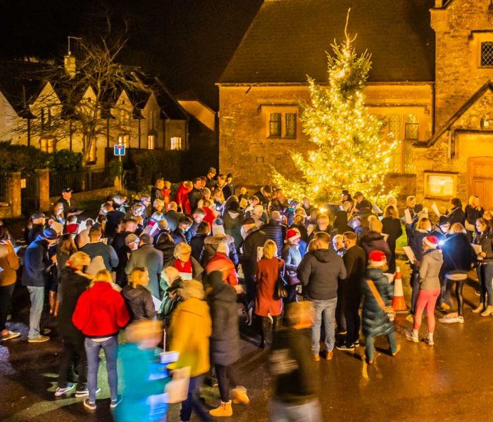 A large group of people sing carols around a lit Christmas tree outside at night.