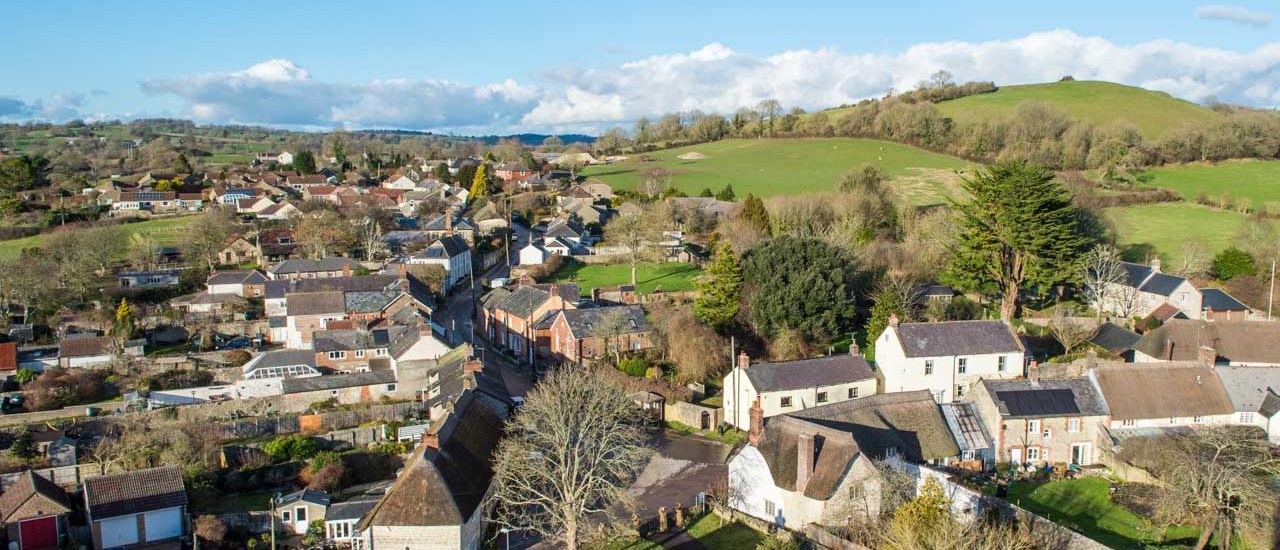 Aerial view of a rural village with houses, trees, and rolling green hills under a blue sky.