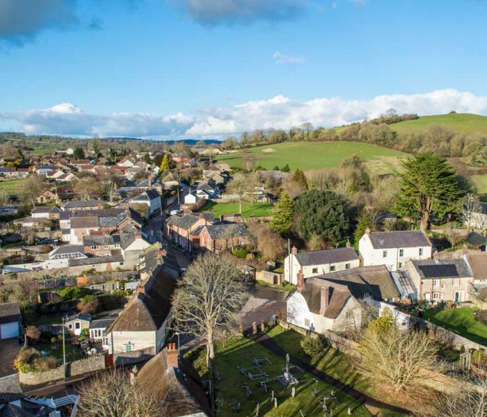 Aerial view of a rural village with houses, trees, and rolling green hills under a blue sky.