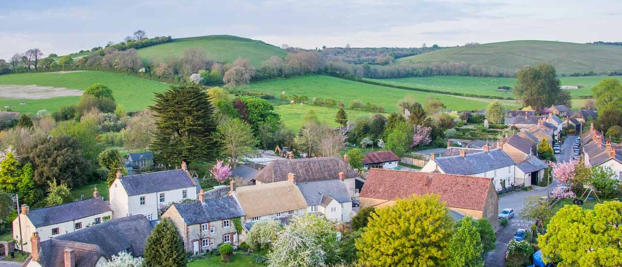 Aerial view of a quaint village with houses, trees, and rolling green hills in the background.