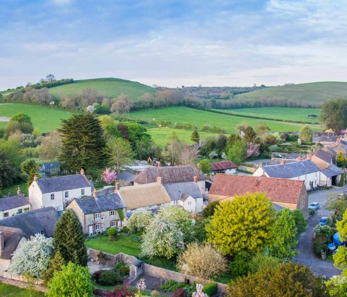 Aerial view of a quaint village with houses, trees, and rolling green hills in the background.